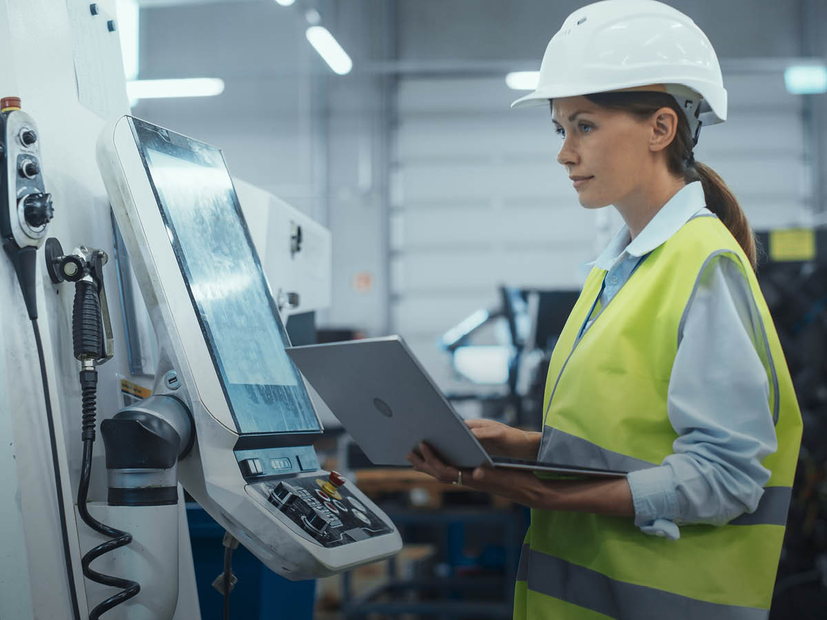 Fachkraft mit Schutzhelm und Warnweste arbeitet mit einem Laptop an einer industriellen Maschine.