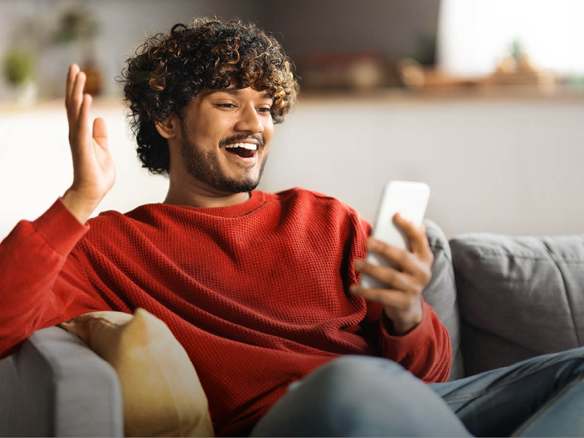 Ein Mann im roten Pulli sitzt auf einem Sofa. Er blickt erfreut in sein Handy, die andere Hand hebt er hoch.