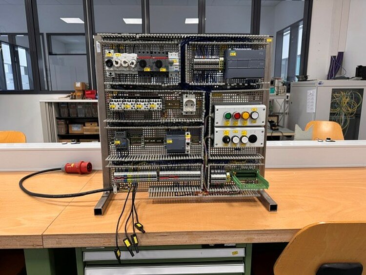 Ein Prüfgestell mit zahlreichen elektrischen Komponenten steht auf einem Arbeitstisch in einem Laborraum. Das Gestell besteht aus mehreren Ebenen mit Schaltern, Steckdosen, Relais, Sicherungen und Steuerungseinheiten. Kabel verlaufen von der unteren Seite des Gestells zum Tisch. Im Hintergrund sind Fenster und Regale mit technischen Geräten zu sehen.