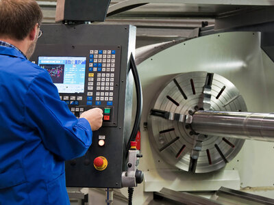 Eine Person in einem blauen Laborkittel bedient eine CNC-Maschine. Die Person arbeitet am Bedienfeld der Maschine, das zahlreiche Tasten, ein Display mit Daten und Grafiken sowie einen Not-Aus-Knopf enthält. In der Maschine ist ein großes zylindrisches Werkstück eingespannt, das offenbar gerade bearbeitet wird.