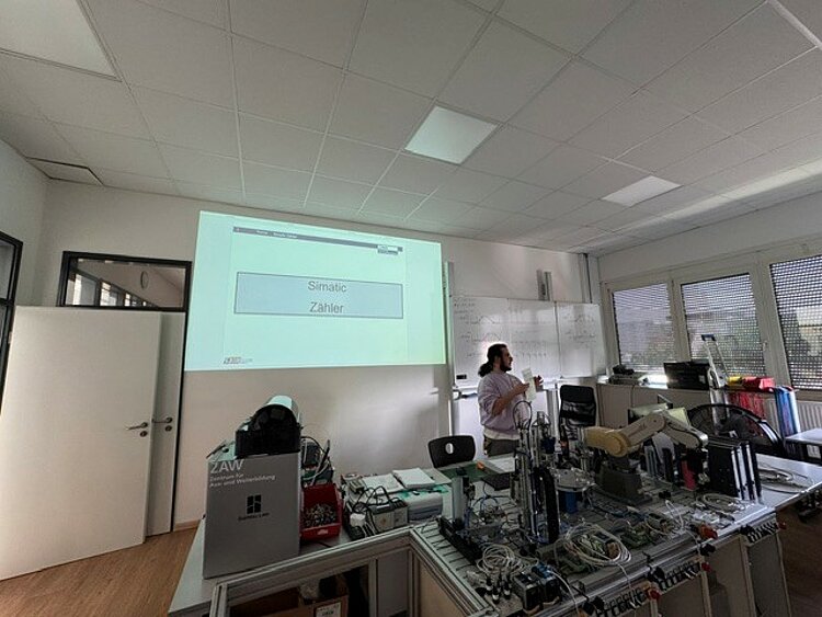 Ein Unterrichtsraum mit mehreren technischen Geräten und Kabeln auf einem Tisch im Vordergrund. Im Hintergrund steht eine Lehrkraft seitlich vor einer weißen Wand und hält einen Zeigestab. An der Wand ist eine große Projektion mit dem Titel 'Simatic Zähler' zu sehen. Die Fenster auf der rechten Seite lassen Tageslicht herein, und an der Wand hängt ein großes Whiteboard mit Notizen.