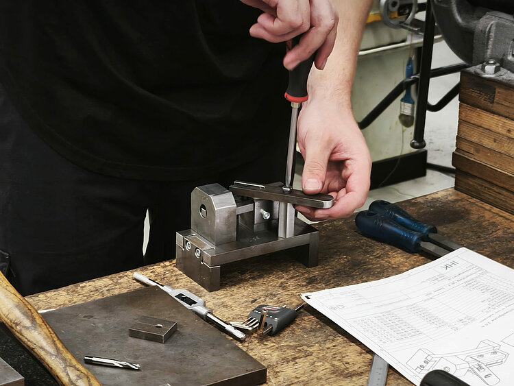 Nahaufnahme einer Person, die an einer Werkbank mit einem Schraubendreher eine Metallvorrichtung bearbeitet. Daneben liegen Werkzeuge, Metallteile und eine technische Zeichnung.
