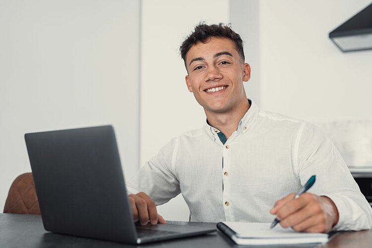 Ein junger Mann mit einem freundlichen Lächeln sitzt an einem Schreibtisch. Er trägt ein weißes Hemd und arbeitet an einem Laptop, während er Notizen in ein Notizbuch schreibt. Auf dem Tisch liegen ein Stift und ein Smartphone. Der Hintergrund ist hell und neutral, was eine konzentrierte Arbeitsatmosphäre vermittelt.