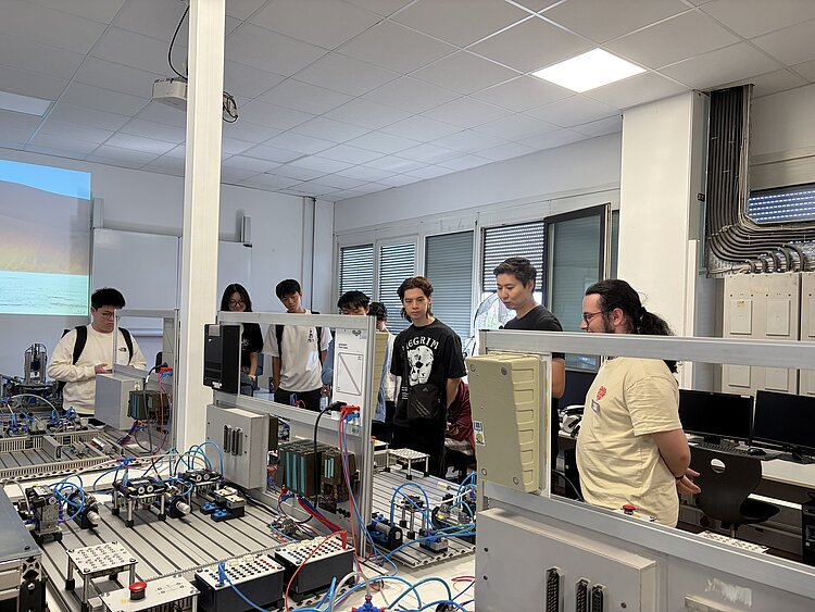 Eine Gruppe junger Menschen besichtigt einen technischen Schulungsraum mit pneumatischen und automatisierten Anlagen. Ein Mitarbeiter erklärt die verschiedenen Steuerungs- und Fertigungssysteme.
