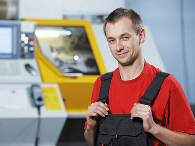 Ein Arbeiter in roter Arbeitskleidung und schwarzer Latzhose steht vor einer gelben CNC-Maschine in einer industriellen Umgebung. Er hält die Träger seiner Latzhose mit beiden Händen fest und lächelt in die Kamera.