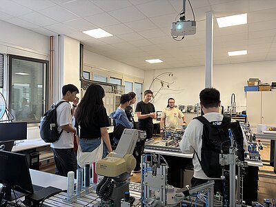 Eine Gruppe informiert sich in einem Automatisierungs- und Robotiklabor über technische Anlagen. Im Vordergrund sind ein Industrieroboter und verschiedene pneumatische Trainingssysteme zu sehen.