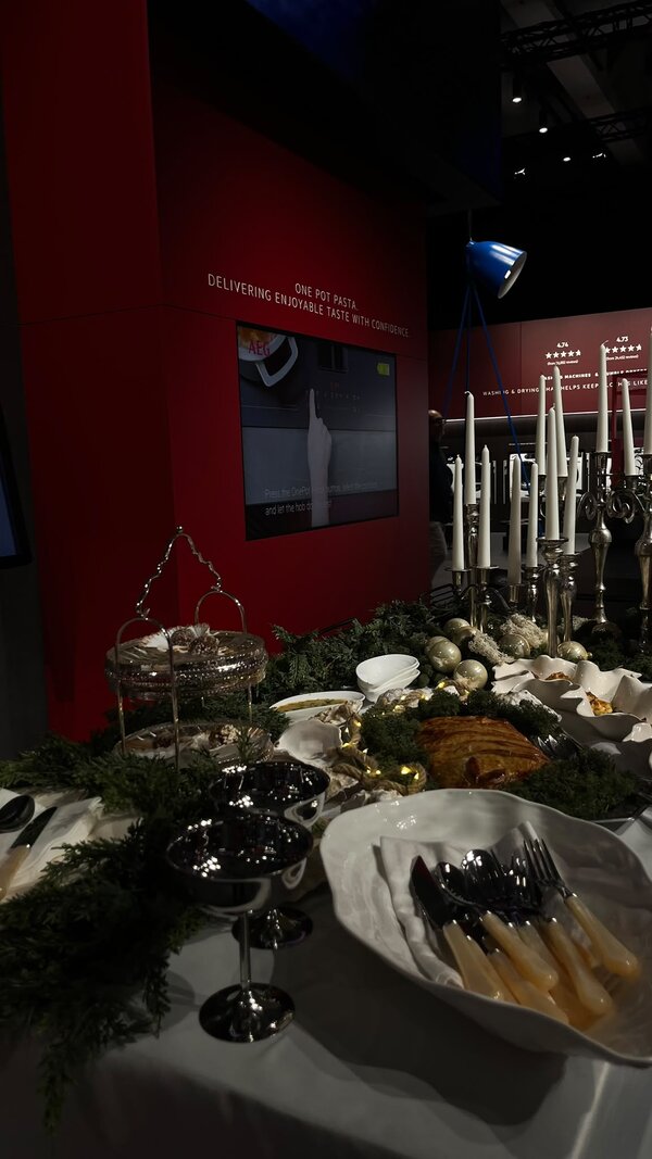 Festlich gedecktes Buffet mit Geschirr, Besteck, Kerzen und Speisen vor einer dunkelroten Präsentationswand in einem Ausstellungsbereich.