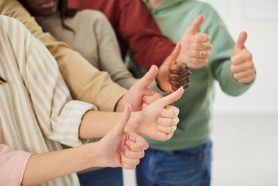 Mehrere Menschen stehen nebeneinander und strecken ihre Arme nach vorne, wobei sie den Daumen nach oben zeigen. Die Kleidung ist unterschiedlich, darunter helle und dunkle Langarmoberteile. Die Szene ist hell ausgeleuchtet, der Hintergrund bleibt unscharf.