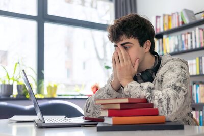 Ein junger Mann sitzt angespannt an einem Schreibtisch in einem hellen Raum mit großen Fenstern. Er hat die Hände vor dem Gesicht gefaltet und stützt die Ellbogen auf einem Stapel bunter Bücher ab. Vor ihm liegt ein offener Laptop, und im Hintergrund sind Regale mit weiteren Büchern und Pflanzen zu sehen. Er trägt einen hellen Strickpullover und Kopfhörer um den Hals.