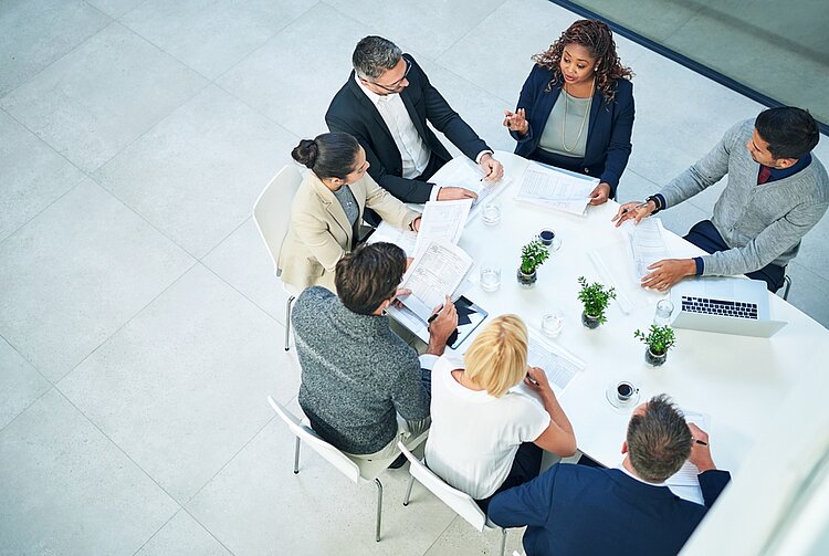 Draufsicht auf eine Gruppe Menschen an einem weißen Besprechungstisch. Die Menschen sprechen miteinander. Auf dem Tisch liegen Papiere und ein Laptop, dazwischen stehen Gläser, Kaffeetassen und Pflanzen.