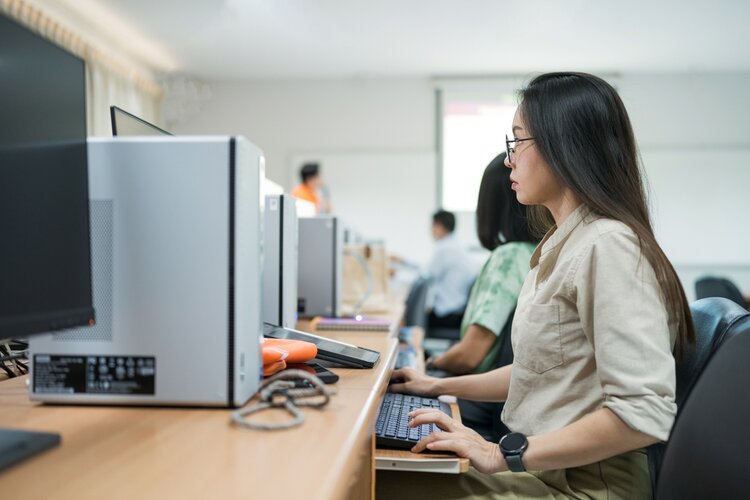 Eine Frau sitzt an einem Schreibtisch vor einem Computer. Sie trägt eine Brille und konzentriert sich auf ihre Arbeit. Der Raum ist voll mit anderen Leuten, die an ihren Computern arbeiten.