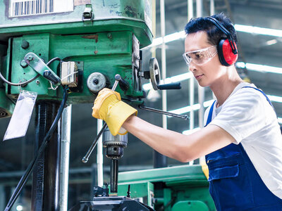 Eine Person bedient eine grüne industrielle Bohrmaschine in einer Werkstatt- oder Fabrikumgebung. Sie trägt gelbe Handschuhe, roten Gehörschutz und einen blauen Overall über einem weißen Hemd. Das Gesicht der Person ist aus Datenschutzgründen unscharf.