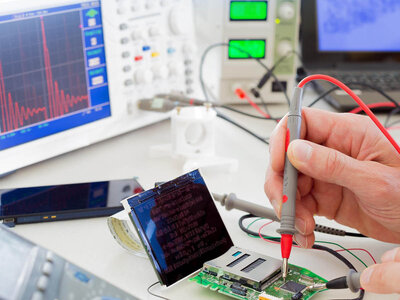 Eine Person verwendet ein Multimeter, um Komponenten auf einer elektronischen Leiterplatte zu testen oder zu messen. Die Umgebung umfasst verschiedene elektronische Geräte wie ein Oszilloskop mit sichtbaren Wellenformen, ein Netzgerät mit digitalen Anzeigen und weitere typische Werkzeuge eines Elektroniklabors.
