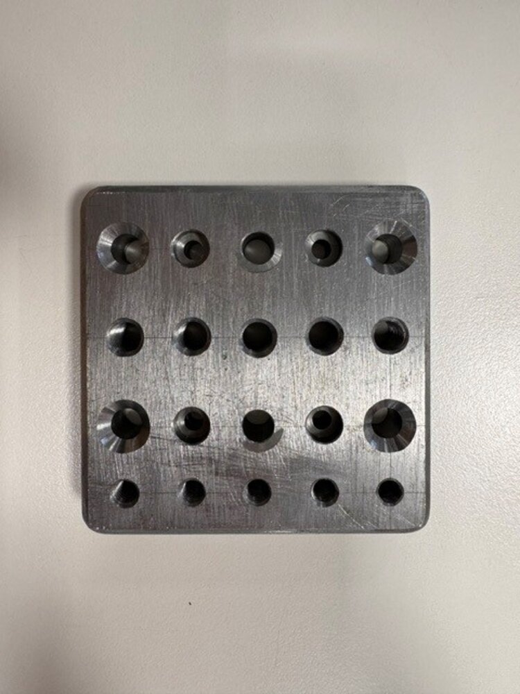 Quadratische Metallplatte mit abgerundeten Ecken und mehreren unterschiedlich großen Bohrungen, die in vier Reihen angeordnet sind. Einige der Bohrungen sind deutlich angesenkt.