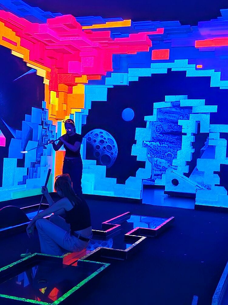 Ein futuristischer, pixeliger Raum mit leuchtenden blauen, pinken und gelben Wänden, die einen Blick ins Weltall durch Maueröffnungen darstellen wie in einem Computerspiel. In dem Raum ist eine Minigolfanlage aufgebaut. Eine Person steht und hält einen Golfschläger, eine andere sitzt auf dem Boden.