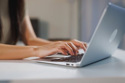 Eine weibliche Person tippt mit beiden Händen auf die Tastatur eines silberfarbenen Laptops, der auf einem weißen Tisch steht. Die Ärmel sind kurz, der Hintergrund ist unscharf und zeigt neutrale Farben.