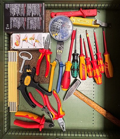 Geöffnete Werkzeugschublade mit verschiedenen Handwerkzeugen, darunter Schraubendreher, Zangen, Hammer, Zollstock, Abisolierwerkzeug und weitere Elektro-Werkzeuge.
