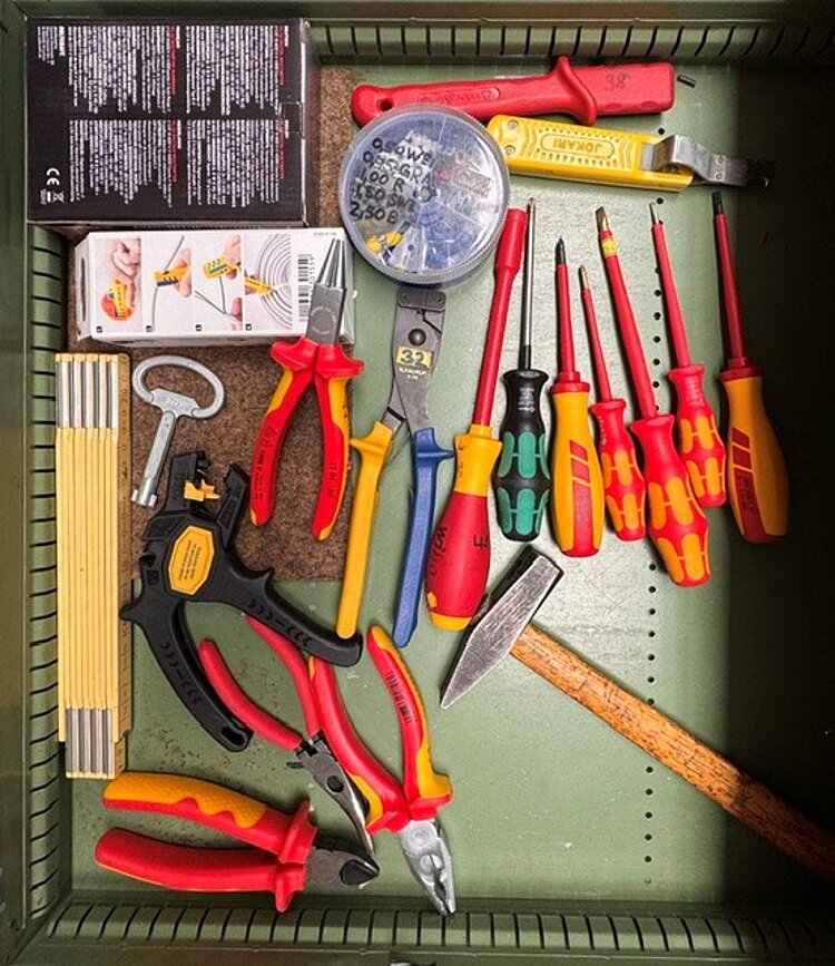 Geöffnete Werkzeugschublade mit verschiedenen Handwerkzeugen, darunter Schraubendreher, Zangen, Hammer, Zollstock, Abisolierwerkzeug und weitere Elektro-Werkzeuge.