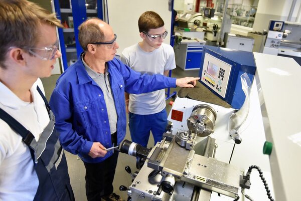 Zwei Auszubildende und ein Ausbilder in Arbeitskleidung und Sicherheitsbrillen beraten sich an einer Drehmaschine in einer Werkstatt.