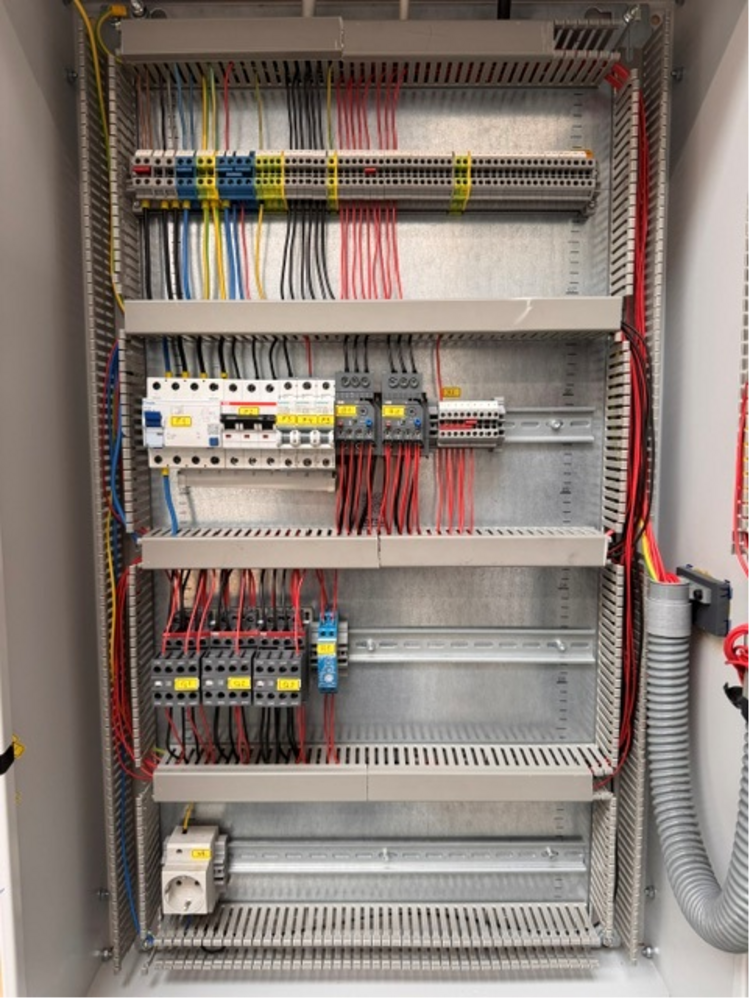 Offener Schaltschrank mit mehreren waagerechten Metallträgern, auf denen verschiedene elektrische Komponenten montiert sind. Mehrere Reihen von Reihenklemmen, Sicherungsautomaten und Relais sind sichtbar. Zahlreiche rote, blaue, gelbe und schwarze Kabel verlaufen geordnet und gebündelt zwischen den Komponenten. Seitlich verlaufen Kabelkanäle und Wellrohre, die die Leitungen aufnehmen. Die Verdrahtung ist übersichtlich und strukturiert ausgeführt.