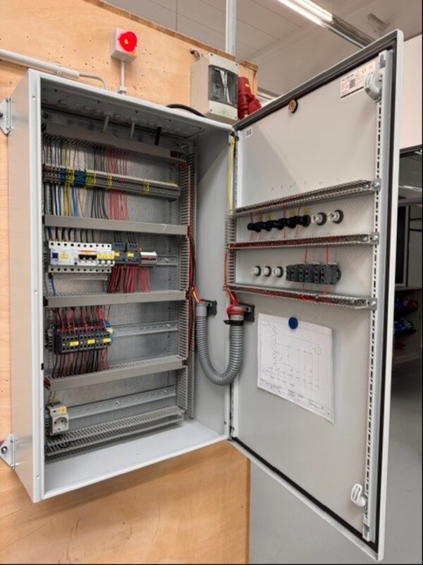 Geöffneter Schaltschrank mit zahlreichen Kabeln, Reihenklemmen, Sicherungsautomaten und Schaltgeräten. Die Verdrahtung ist ordentlich geführt und farblich sortiert. Auf der Innenseite der Tür ist ein Schaltplan befestigt. Das Gehäuse steht vor einer hellen Wand, oben ist ein roter Signalgeber montiert.