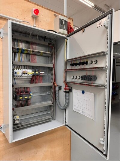 Geöffneter Schaltschrank mit zahlreichen Kabeln, Reihenklemmen, Sicherungsautomaten und Schaltgeräten. Die Verdrahtung ist ordentlich geführt und farblich sortiert. Auf der Innenseite der Tür ist ein Schaltplan befestigt. Das Gehäuse steht vor einer hellen Wand, oben ist ein roter Signalgeber montiert.