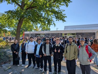 Eine Gruppe junger Teilnehmender steht im Vordergrund auf einem gepflasterten Weg im Freien. Sie tragen überwiegend sportliche oder legere Kleidung, einige haben Rucksäcke dabei. Im Hintergrund sind weitere Menschen vor einem flachen Gebäude mit großen Fenstern zu sehen. Links ragt ein großer Baum mit ausladender Krone ins Bild, der Schatten auf den Weg wirft. Die Szene wirkt wie eine Versammlung oder ein Treffen bei sonnigem Wetter.