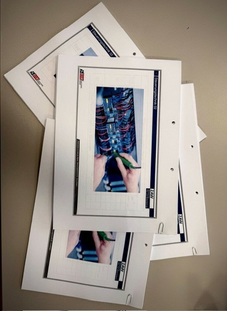 Mehrere Arbeitsblätter zur Elektro- oder Verdrahtungstechnik liegen auf einem Tisch; auf dem obersten Blatt ist ein Foto von Arbeiten an einem Schaltschrank zu sehen.