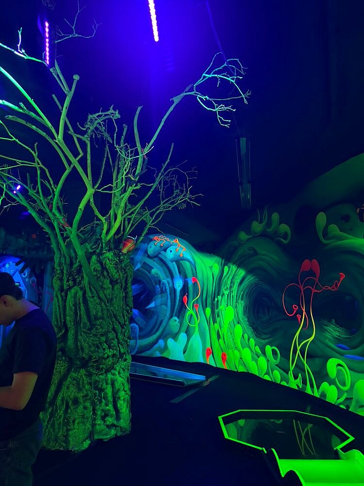 Ein dunkler Raum mit leuchtenden, grünen und blauen Neonfarben, der eine Unterwasserwelt darstellt. Im Vordergrund steht ein künstlicher Baum mit leuchtenden Ästen, während im Hintergrund eine Wand mit Meeresmotiven wie Quallen und Korallen zu sehen ist.