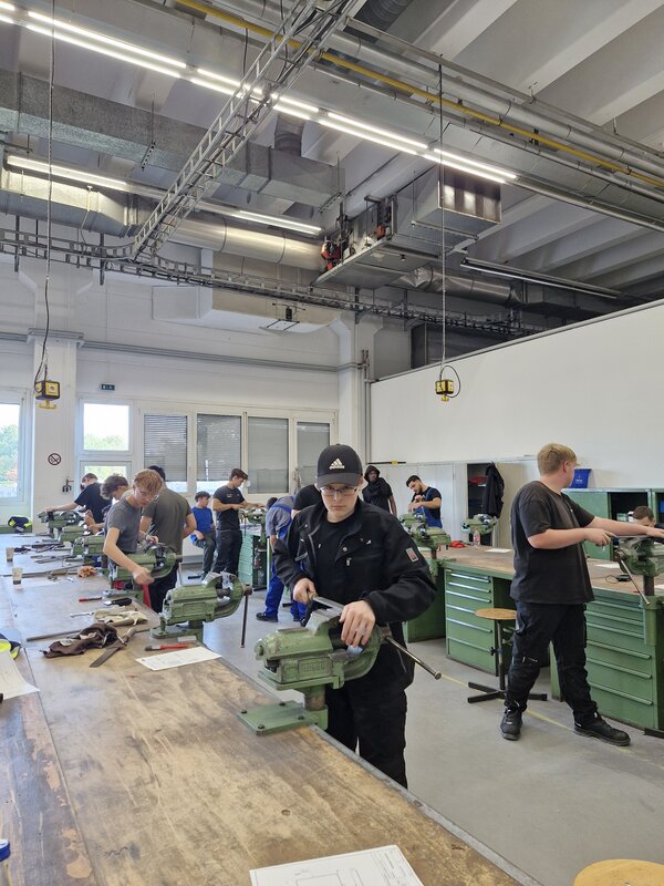 Auszubildende arbeiten in einer großen Metallwerkstatt an Werkbänken und Schraubstöcken; im Hintergrund stehen zahlreiche Maschinen und Werkzeugschränke.