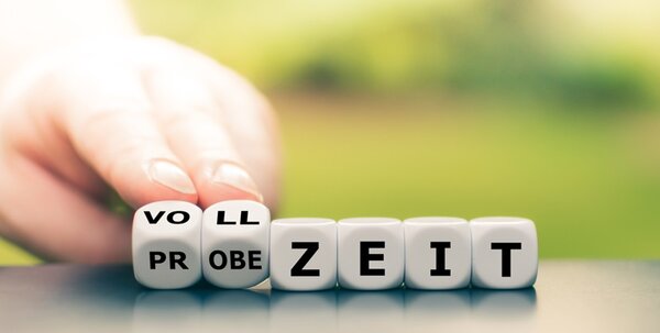 Im Vordergrund sind sechs Würfel mit Buchstaben zu sehen, die das Wort 'PROBEZEIT' und teilweise 'VOLLZEIT' bilden. Eine Hand berührt die Würfel von links. Der Hintergrund ist unscharf und zeigt grüne Farbtöne.