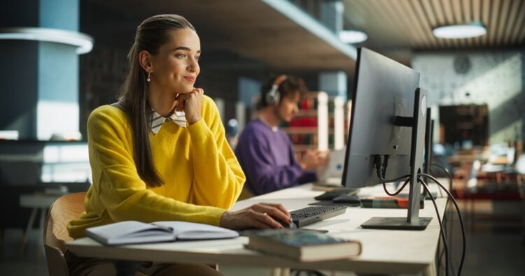 Eine junge Frau mit langen, dunklen Haaren sitzt an einem Schreibtisch und arbeitet am Computer. Sie trägt einen gelben Pullover und eine weiße Bluse. Ihr Blick ist konzentriert auf den Bildschirm gerichtet, die rechte Hand liegt auf der Maus. Auf dem Tisch befinden sich ein aufgeschlagenes Notizbuch, ein Stift und ein Buch. Im Hintergrund sitzt eine weitere Person an einem Computer, die Kopfhörer trägt. Die Umgebung wirkt modern und hell, mit großen Fenstern und einer Deckenstruktur aus Holzlamellen.