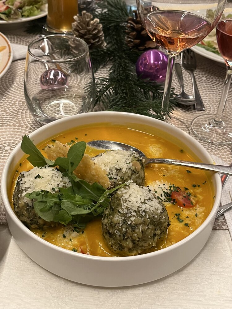 Eine Teller mit drei Spinatknödeln, etwas Salat und Parmesan in einer gelben Sauce. Hinter dem Teller befinden sich verschiedene Gläser und weihnachtliche Dekoration.