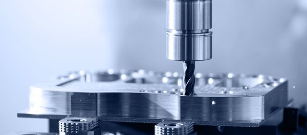 Nahaufnahme einer Fräsmaschine, die mit einem rotierenden Fräser ein Metallwerkstück bearbeitet. Dabei sind feine Metallspäne rund um die Bohrstelle zu sehen.