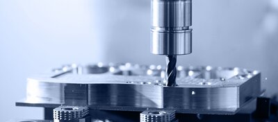 Nahaufnahme einer Fräsmaschine, die mit einem rotierenden Fräser ein Metallwerkstück bearbeitet. Dabei sind feine Metallspäne rund um die Bohrstelle zu sehen.