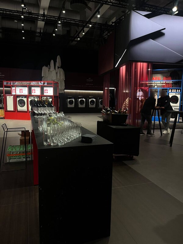 Blick in einen modern gestalteten Ausstellungsbereich für Haushaltsgeräte mit Waschmaschinen, Präsentationsflächen und bereitgestellten Gläsern im Vordergrund.