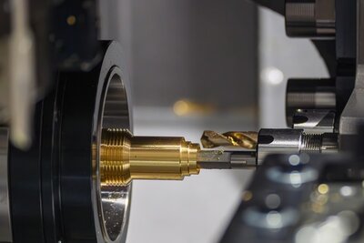 Im Vordergrund befindet sich ein goldfarbenes Metallteil, das in eine Maschine eingespannt ist. Ein Werkzeug mit silbernem Schaft und goldener Spitze bearbeitet das Metallteil präzise. Die Szene zeigt eine industrielle Umgebung mit klaren, glänzenden Oberflächen und mechanischen Komponenten.