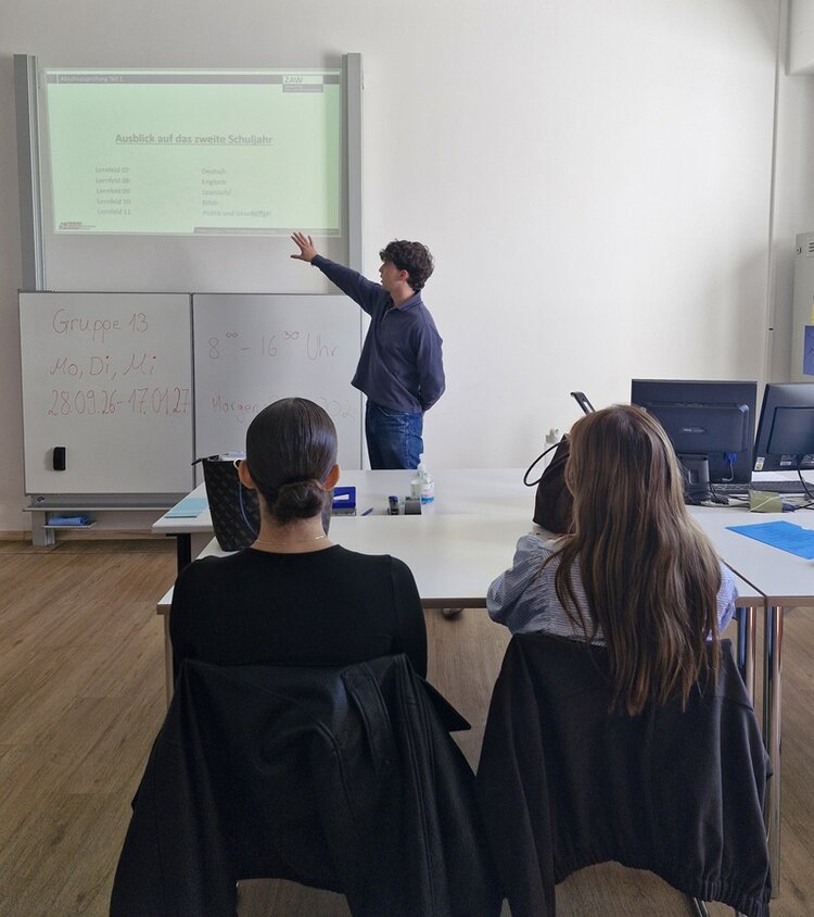 Ein Auszubildender steht vor einer Projektionsfläche und zeigt mit ausgestrecktem Arm auf einen Text, der oben auf der Fläche zu sehen ist. Zwei Auszubildende sitzen mit dem Rücken zur Kamera an einem Tisch und schauen in Richtung ihres Mitazubis. Auf einem Whiteboard neben der Projektionsfläche sind handschriftliche Notizen zu sehen. Der Raum wirkt hell und modern, mit weißen Wänden und Holzfußboden.