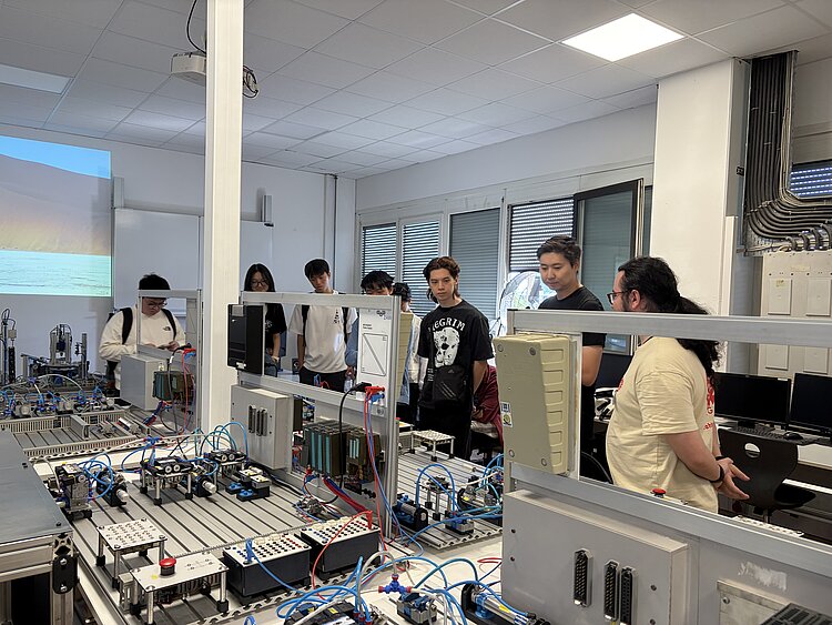 Junge Besucher*innen stehen gemeinsam mit einem Mitarbeiter vor einer komplexen automatisierten Trainingsanlage mit Steuerungen, Sensoren und Drucklufttechnik.