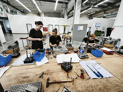 Drei junge Erwachsene arbeiten gemeinsam an einer großen Werkbank in einer Metallwerkstatt. Vor ihnen liegen Schraubstöcke, Bohrer, Feilen, Messwerkzeuge und technische Zeichnungen.