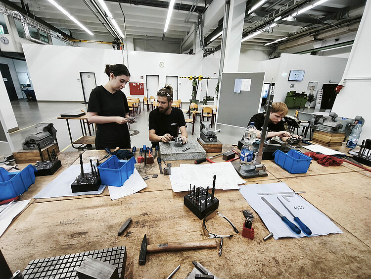 Drei junge Erwachsene arbeiten gemeinsam an einer großen Werkbank in einer Metallwerkstatt. Vor ihnen liegen Schraubstöcke, Bohrer, Feilen, Messwerkzeuge und technische Zeichnungen.