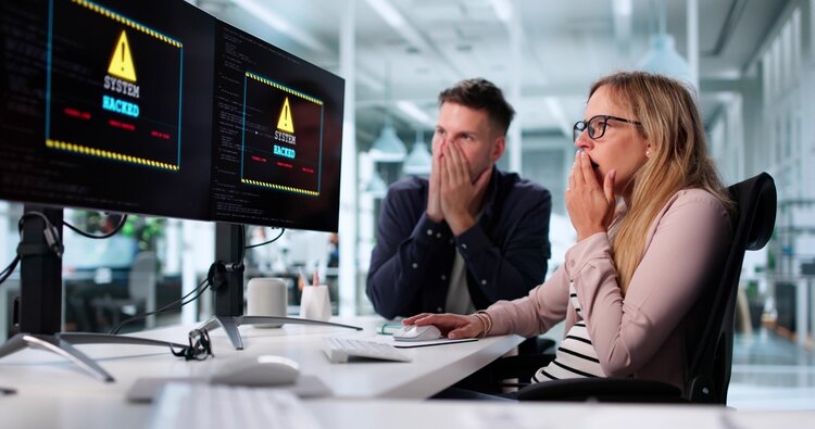 Cyberangriff auf einen Geschäftscomputer: Ransomware Malware bedroht Dateien und viele Alarmzeichen sind auf einem Bildschirm zu sehen. Zwei Mitarbeiter*innen sitzen mit Hand vor dem Mund davor.