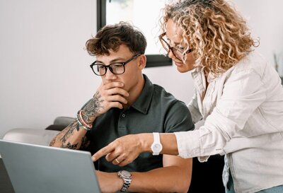 Eine Frau mit blonden Locken und einer Brille zeigt einem jungen Mann mit Tattoos und einer Brille etwas auf einem Laptop, während der junge Mann in einer nachdenklichen Geste seine Hand an seinen Mund hält.