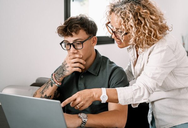 Eine Frau mit blonden Locken und einer Brille zeigt einem jungen Mann mit Tattoos und einer Brille etwas auf einem Laptop, während der junge Mann in einer nachdenklichen Geste seine Hand an seinen Mund hält.