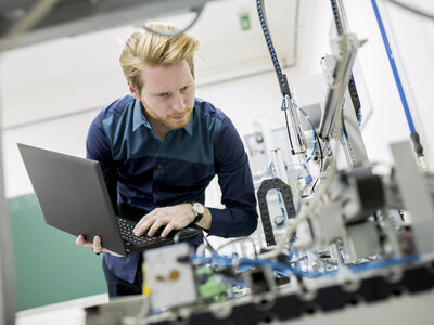 Eine Person arbeitet mit einem Laptop in einer industriellen oder laborähnlichen Umgebung. Um sie herum befinden sich verschiedene Maschinen und Geräte, möglicherweise im Bereich Automatisierung oder Robotik. Das Gesicht der Person ist aus Datenschutzgründen unscharf.