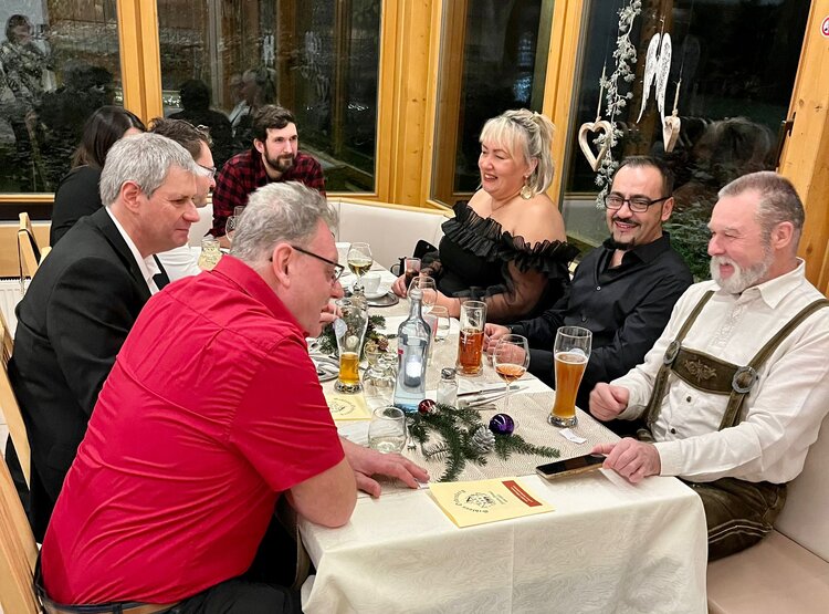 Acht Personen sitzen an einem gedeckten Tisch im Wintergarten eines Restaurants. Draußen ist es dunkel und die acht Personen unterhalten sich und lachen miteinander.