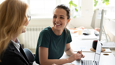 Zwei junge Frauen sitzen an einem Schreibtisch und lächeln sich an.