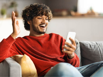 Ein Mann im roten Pulli sitzt auf einem Sofa. Er blickt erfreut in sein Handy, die andere Hand hebt er hoch.