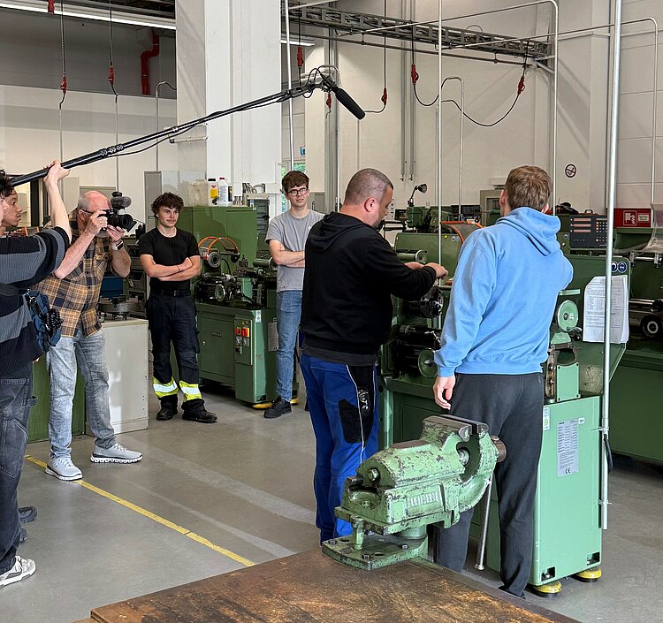 Filmteam bei einem Dreh in einer Maschinenhalle. Ein Kameramann und ein Tontechniker begleiten eine Szene zwischen Werkzeugmaschinen und jungen Männern.