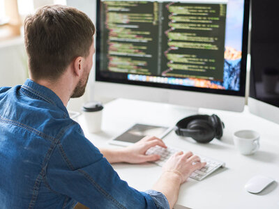 Eine Person sitzt an einem Schreibtisch und arbeitet an einem Computer. Auf dem Bildschirm sind Codezeilen zu sehen. Auf dem Schreibtisch befinden sich außerdem eine Kaffeetasse, Kopfhörer und ein Tablet.